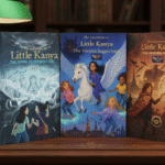 A set of three hardcover adventure books from 'The Little Kanya Trilogy' displayed on a wooden desk. The titles are 'The Sonic Reverberator' in blue, 'The Vienna Juggernaut' featuring a girl on a winged horse, and 'The Moscow Mosaic' in warm orange tones. A vintage green library lamp and a cup of tea sit in the background.